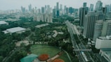 A sprawling urban landscape featuring numerous high-rise buildings with various modern architectural designs. A major roadway cuts through the cityscape, flanked by lush greenery including parks and sports fields, creating a blend of nature and urban development.