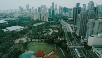 A sprawling urban landscape featuring numerous high-rise buildings with various modern architectural designs. A major roadway cuts through the cityscape, flanked by lush greenery including parks and sports fields, creating a blend of nature and urban development.