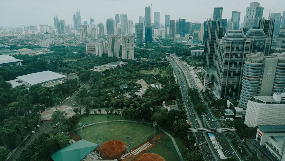 A sprawling urban landscape featuring numerous high-rise buildings with various modern architectural designs. A major roadway cuts through the cityscape, flanked by lush greenery including parks and sports fields, creating a blend of nature and urban development.