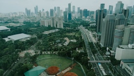 A sprawling urban landscape featuring numerous high-rise buildings with various modern architectural designs. A major roadway cuts through the cityscape, flanked by lush greenery including parks and sports fields, creating a blend of nature and urban development.