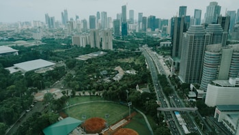A sprawling urban landscape featuring numerous high-rise buildings with various modern architectural designs. A major roadway cuts through the cityscape, flanked by lush greenery including parks and sports fields, creating a blend of nature and urban development.