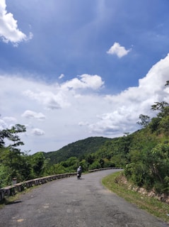 A winding road curves through a green, hilly landscape under a bright blue sky with scattered white clouds. A person wearing a helmet rides a motorcycle along the road, surrounded by lush foliage. The scene exudes a sense of tranquility and openness.