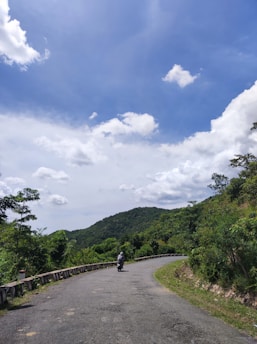 A winding road curves through a green, hilly landscape under a bright blue sky with scattered white clouds. A person wearing a helmet rides a motorcycle along the road, surrounded by lush foliage. The scene exudes a sense of tranquility and openness.