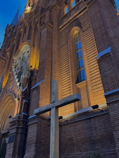 The image depicts a large, ornate brick building with intricate architectural details and a tall cross positioned in the foreground. The building features arched windows and decorative carvings, illuminated by warm lighting that highlights its textures against a blue evening sky.