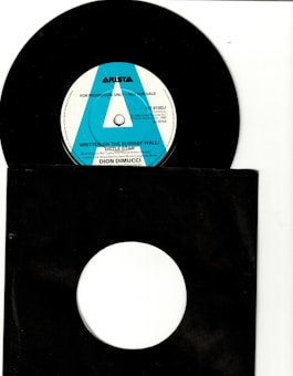A black vinyl record with a white and blue label in the center is partially visible. The label includes text such as 'ARISTA' and 'FOR PROMOTION ONLY - NOT FOR SALE.' The record is placed inside a black sleeve with a circular cutout, revealing the label. The surrounding background is mostly white.