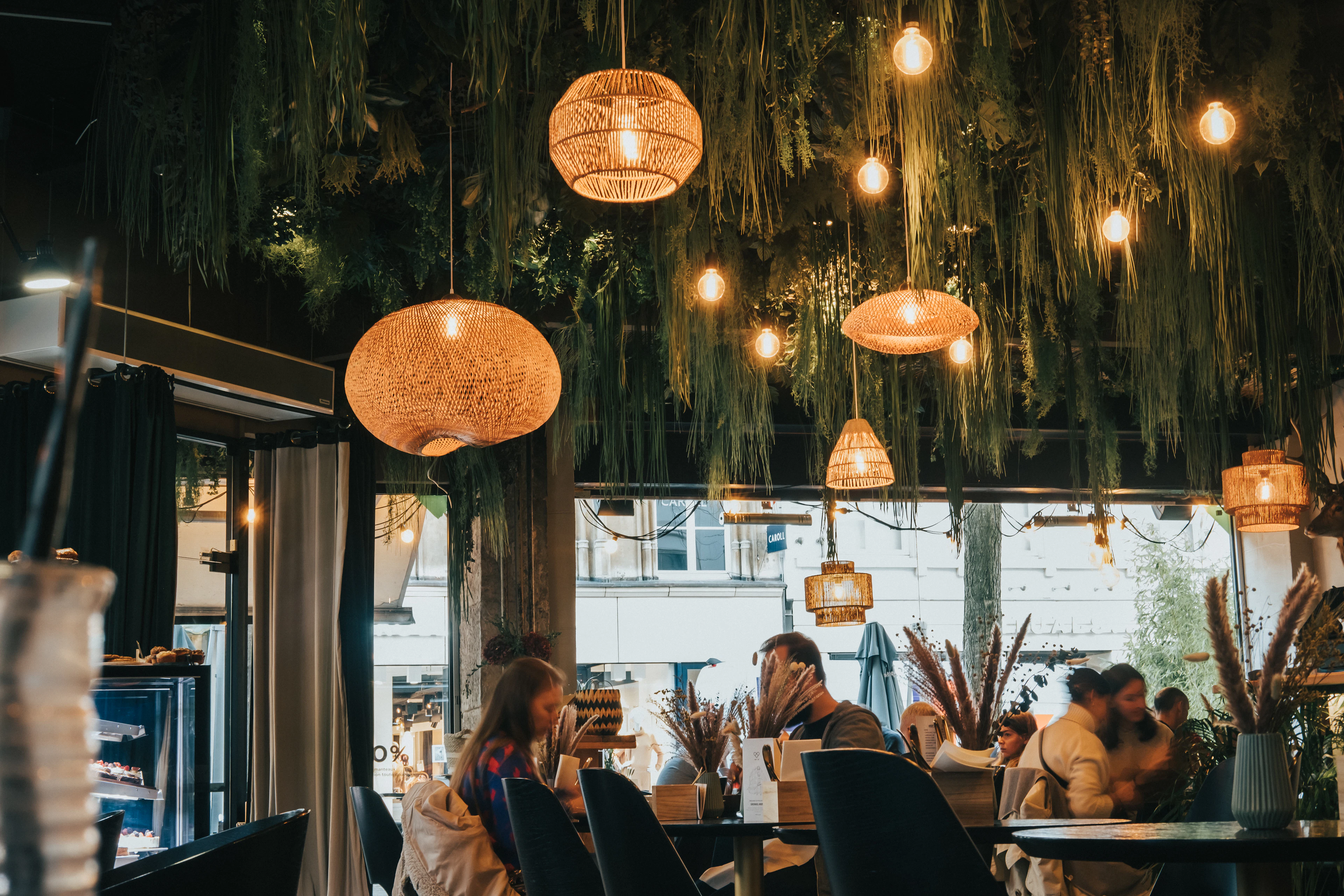 Diners sit beneath hanging woven lampshades and greenery inside a warmly lit restaurant.
