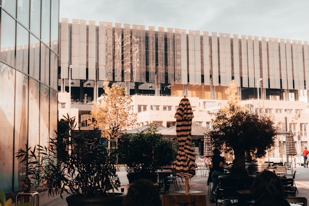 An urban outdoor setting featuring a modern building with large, dark reflective windows in the background. In the foreground, there are potted plants and outdoor seating which includes tables and chairs. Natural elements such as small trees and shrubs add greenery to the area. An umbrella with stripes is closed and positioned among the seating.