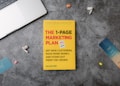 A copy of the book titled 'The 1-Page Marketing Plan' lies on a gray textured surface. The book cover is bright yellow with bold red, white, and green text. Surrounding the book are various small objects: a metallic laptop partially visible on the top left, a pair of white wireless earphones, a blue bookmark with the text 'one more page,' and other small, colorful items.