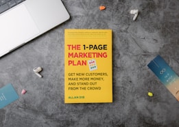 A copy of the book titled 'The 1-Page Marketing Plan' lies on a gray textured surface. The book cover is bright yellow with bold red, white, and green text. Surrounding the book are various small objects: a metallic laptop partially visible on the top left, a pair of white wireless earphones, a blue bookmark with the text 'one more page,' and other small, colorful items.