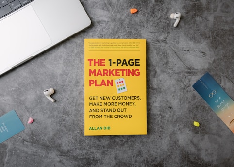 A copy of the book titled 'The 1-Page Marketing Plan' lies on a gray textured surface. The book cover is bright yellow with bold red, white, and green text. Surrounding the book are various small objects: a metallic laptop partially visible on the top left, a pair of white wireless earphones, a blue bookmark with the text 'one more page,' and other small, colorful items.