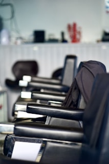 A row of black salon chairs arranged in a hair salon, with a jacket draped over one of the chairs. The background features blurred objects, possibly hair products or equipment.