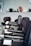 A row of black salon chairs arranged in a hair salon, with a jacket draped over one of the chairs. The background features blurred objects, possibly hair products or equipment.