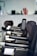 A row of black salon chairs arranged in a hair salon, with a jacket draped over one of the chairs. The background features blurred objects, possibly hair products or equipment.
