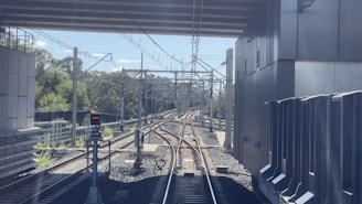 A railway track extends forward under a bridge, flanked by steel railings and electric poles. Vegetation can be seen on the left, along with a signal light that is currently red. On the right is a large industrial-looking building with metal panels.
