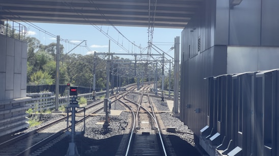 A railway track extends forward under a bridge, flanked by steel railings and electric poles. Vegetation can be seen on the left, along with a signal light that is currently red. On the right is a large industrial-looking building with metal panels.