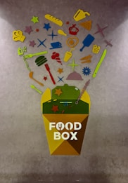 A colorful abstract art piece featuring various food-related items such as a chef's hat, baguette, pizza slice, utensils, and a cup, arranged above a container labeled 'Food Box'. These items are depicted in bright colors including yellow, red, green, and blue, and are against a textured gray background.
