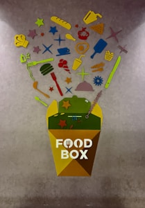 A colorful abstract art piece featuring various food-related items such as a chef's hat, baguette, pizza slice, utensils, and a cup, arranged above a container labeled 'Food Box'. These items are depicted in bright colors including yellow, red, green, and blue, and are against a textured gray background.