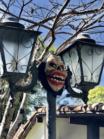 A spooky mask with exaggerated features is affixed to a lamppost between two vintage street lamps. Bare tree branches form a backdrop, creating a contrast with the bright blue sky. In the background, part of a house with a tiled roof is partially visible.