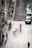 A city street scene with people walking along a tiled pedestrian area. The motion blur of the pedestrians suggests a sense of movement and busyness. Cars are parked along the side of a narrow road, and several buildings line the street, adding an urban architectural backdrop.