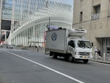 A delivery truck is driving on a city street with a modern architectural building in the background. The building features a distinctive white ribbed facade. Traffic is sparse, and a few pedestrians are visible on the sidewalk.