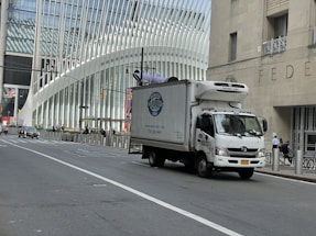 A delivery truck is driving on a city street with a modern architectural building in the background. The building features a distinctive white ribbed facade. Traffic is sparse, and a few pedestrians are visible on the sidewalk.