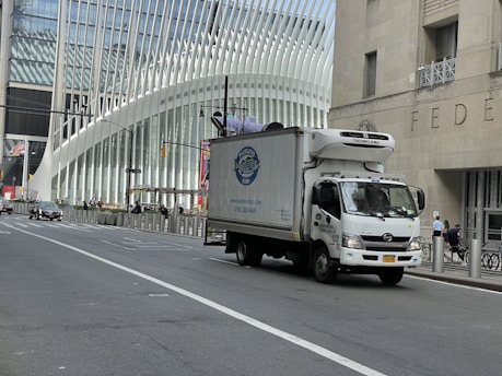 A delivery truck is driving on a city street with a modern architectural building in the background. The building features a distinctive white ribbed facade. Traffic is sparse, and a few pedestrians are visible on the sidewalk.