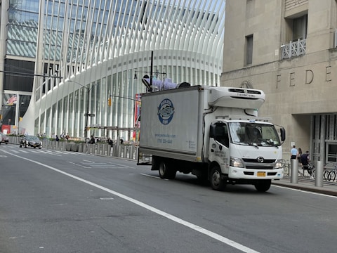 A delivery truck is driving on a city street with a modern architectural building in the background. The building features a distinctive white ribbed facade. Traffic is sparse, and a few pedestrians are visible on the sidewalk.