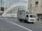 A delivery truck is driving on a city street with a modern architectural building in the background. The building features a distinctive white ribbed facade. Traffic is sparse, and a few pedestrians are visible on the sidewalk.