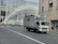 A delivery truck is driving on a city street with a modern architectural building in the background. The building features a distinctive white ribbed facade. Traffic is sparse, and a few pedestrians are visible on the sidewalk.