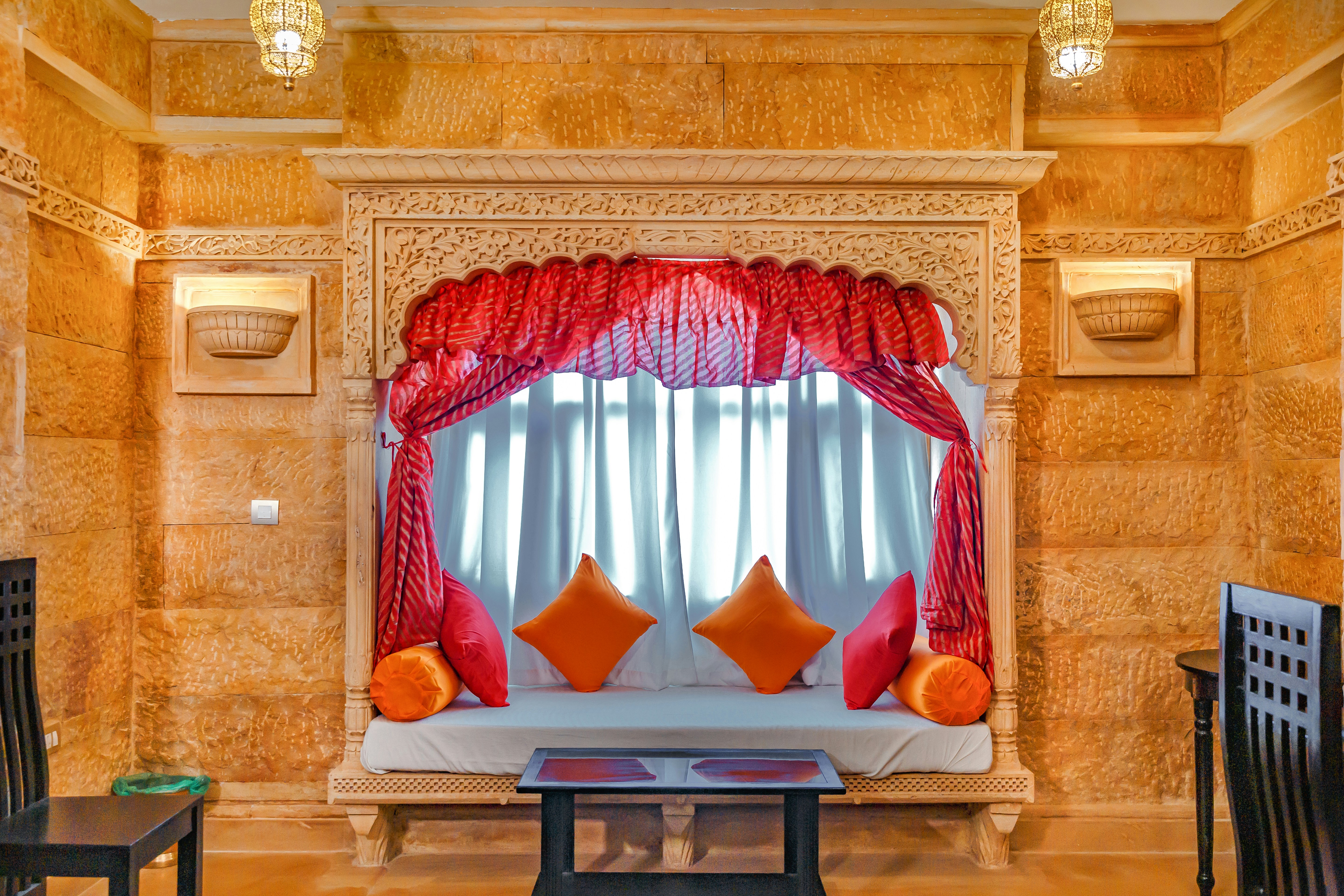 a couch with pillows and a table, Plunge into the unparalleled sense of comfort stay at our Hotel! When visiting the desert city of Jaisalmer, staying at our luxurious hotel is a must to do. Hotel Lal Garh Fort and Palace will add an additional comfort, royalness and luxuriousness to your trip. Enjoy a fort view while sitting at the rooftop cafe and having your favourite delicious food cooked at the in-house restaurant. We assure you to give an unforgettable trip to Jaisalmer with our comfortable stay and delicious food. Book a comfortable and luxurious stay right now. Call us now at +91 9414150150 or +91-8949198625