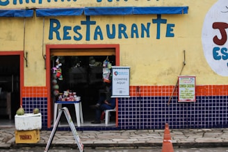 A restaurant entrance with a yellow wall, featuring bright blue letters spelling 'Restaurante'. The lower portion of the wall is decorated with blue and orange tiles. A small table with various goods is visible through the open doorway, and signs are posted next to the entrance, advertising goods and home-cooked meals. A person is sitting inside, partially visible. A yellow crate with two green coconuts and an orange traffic cone are in the foreground.