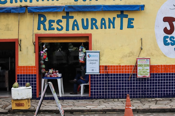 A restaurant entrance with a yellow wall, featuring bright blue letters spelling 'Restaurante'. The lower portion of the wall is decorated with blue and orange tiles. A small table with various goods is visible through the open doorway, and signs are posted next to the entrance, advertising goods and home-cooked meals. A person is sitting inside, partially visible. A yellow crate with two green coconuts and an orange traffic cone are in the foreground.
