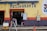 A restaurant entrance with a yellow wall, featuring bright blue letters spelling 'Restaurante'. The lower portion of the wall is decorated with blue and orange tiles. A small table with various goods is visible through the open doorway, and signs are posted next to the entrance, advertising goods and home-cooked meals. A person is sitting inside, partially visible. A yellow crate with two green coconuts and an orange traffic cone are in the foreground.