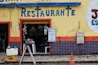 A restaurant entrance with a yellow wall, featuring bright blue letters spelling 'Restaurante'. The lower portion of the wall is decorated with blue and orange tiles. A small table with various goods is visible through the open doorway, and signs are posted next to the entrance, advertising goods and home-cooked meals. A person is sitting inside, partially visible. A yellow crate with two green coconuts and an orange traffic cone are in the foreground.