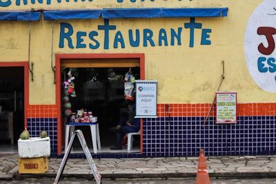 A restaurant entrance with a yellow wall, featuring bright blue letters spelling 'Restaurante'. The lower portion of the wall is decorated with blue and orange tiles. A small table with various goods is visible through the open doorway, and signs are posted next to the entrance, advertising goods and home-cooked meals. A person is sitting inside, partially visible. A yellow crate with two green coconuts and an orange traffic cone are in the foreground.