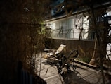 A dimly lit outdoor patio features a wooden deck with a lounge chair and a baby stroller. Bamboo plants on either side create a natural screen, and the background shows a building with large glass windows. A broom leans against the wall near the stroller.