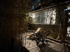 A dimly lit outdoor patio features a wooden deck with a lounge chair and a baby stroller. Bamboo plants on either side create a natural screen, and the background shows a building with large glass windows. A broom leans against the wall near the stroller.