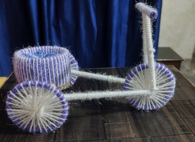 A small, handmade model of a cycle crafted using white and purple yarn. The wheels, handlebars, and seat are wrapped in thick yarn, giving a textured appearance. The backdrop is a deep blue curtain, and the cycle is placed on a dark wooden surface.