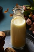 A glass bottle filled with a creamy liquid, possibly milk or a milkshake, is placed on a metallic surface. Surrounding the bottle are some cookies and blurred flowers in the background, creating a cozy and inviting atmosphere.
