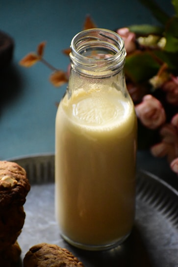 A glass bottle filled with a creamy liquid, possibly milk or a milkshake, is placed on a metallic surface. Surrounding the bottle are some cookies and blurred flowers in the background, creating a cozy and inviting atmosphere.