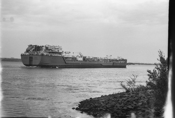 A large cargo ship is navigating through a wide river or a body of water. The vessel features multiple structures on its deck, indicating it is likely used for transporting goods. The sky above is overcast, creating a muted lighting effect. On the right-hand side, there is rocky shoreline with some sparse vegetation.