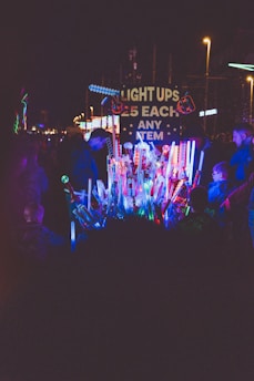 A vibrant night scene with a collection of colorful light-up toys on display, surrounded by people. The sign reads 'Light Ups £5 Each Any Item', hinting at a carnival or fair setting. The warm glow of street lamps and the bright, multicolored lights create a lively atmosphere.