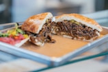 A sandwich filled with shredded, seasoned meat and vegetables sits on a metal tray. The bread looks toasted, and the filling appears juicy with visible herbs. Alongside the sandwich, there is a small portion of a colorful salad, including sliced tomatoes and peppers.