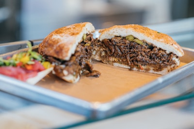 A sandwich filled with shredded, seasoned meat and vegetables sits on a metal tray. The bread looks toasted, and the filling appears juicy with visible herbs. Alongside the sandwich, there is a small portion of a colorful salad, including sliced tomatoes and peppers.