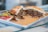 A sandwich filled with shredded, seasoned meat and vegetables sits on a metal tray. The bread looks toasted, and the filling appears juicy with visible herbs. Alongside the sandwich, there is a small portion of a colorful salad, including sliced tomatoes and peppers.