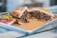 A sandwich filled with shredded, seasoned meat and vegetables sits on a metal tray. The bread looks toasted, and the filling appears juicy with visible herbs. Alongside the sandwich, there is a small portion of a colorful salad, including sliced tomatoes and peppers.