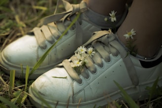 A pair of white sneakers adorned with small white flowers and green stems is set against a backdrop of grass and earth. The shoes have slightly worn laces and a few smudges, giving them a casual, natural appearance. The contrast between the clean white shoes and the delicate flowers highlights a blend of urban and natural elements.