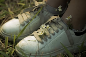 A pair of white sneakers adorned with small white flowers and green stems is set against a backdrop of grass and earth. The shoes have slightly worn laces and a few smudges, giving them a casual, natural appearance. The contrast between the clean white shoes and the delicate flowers highlights a blend of urban and natural elements.