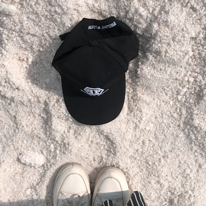 A black baseball cap rests on a textured, granular surface resembling salt or sand. Below the cap, a pair of white shoes with a gray inset and striped clothing are partially visible, suggesting someone standing nearby.