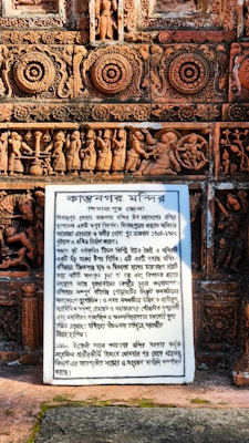 Intricate terracotta carvings adorn a temple wall, featuring floral and geometric patterns as well as detailed figures depicting scenes from mythology. A white plaque with inscriptions provides historical context in a local script.
